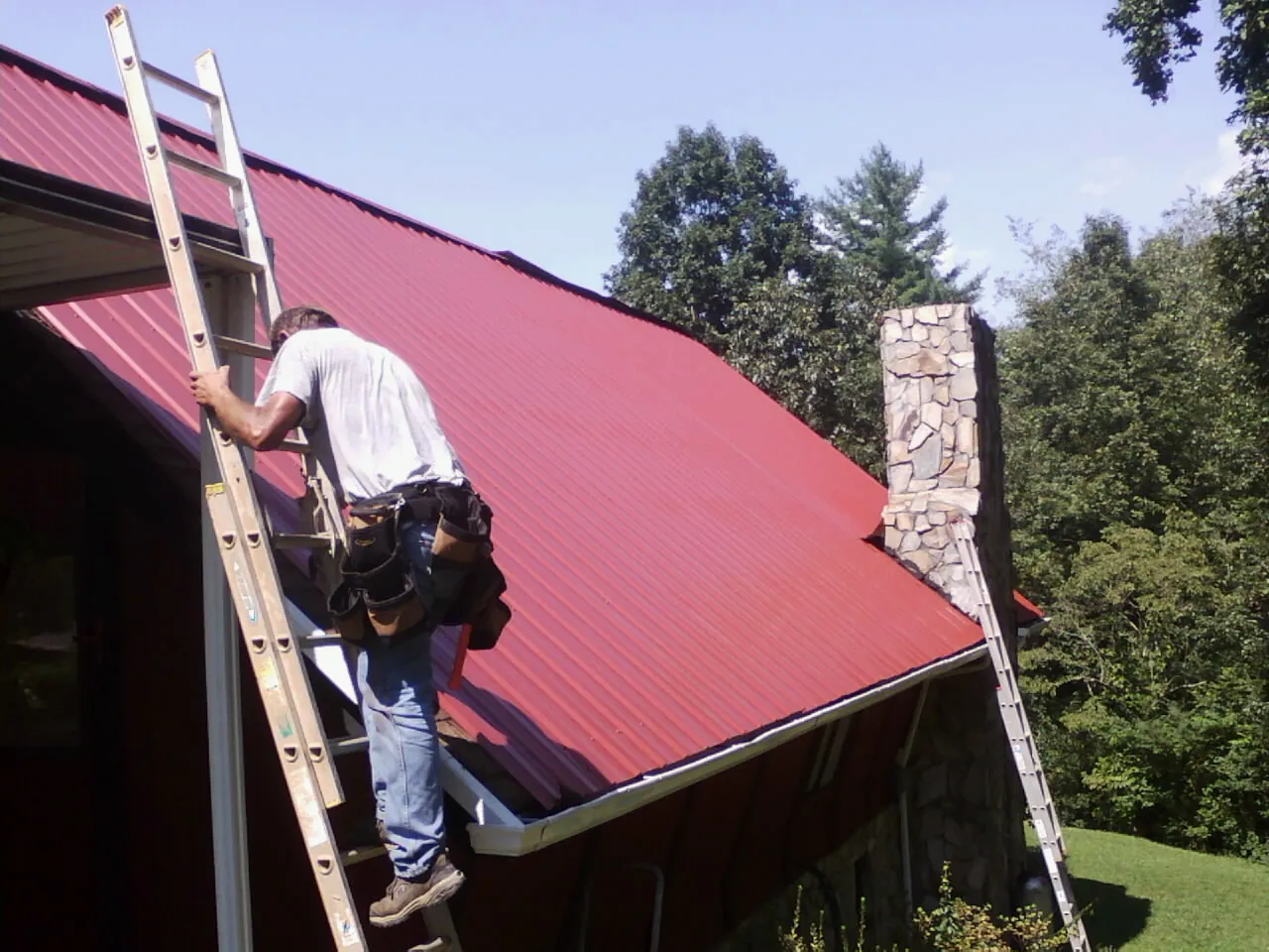 Roofer on a ladder beside a red metal roof and stone chimney — Soffit & Fascia Repair by Roofing Brothers in Pompton Plains